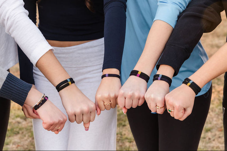 Several people standing outdoors with their hands together, each wearing a black fitness tracker with a colorful band.